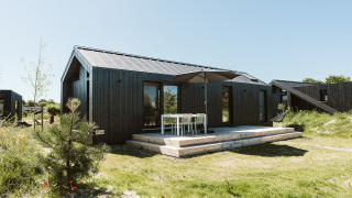 Casa pequeña negra con terraza de madera y muebles de exterior en De Klepperstee, Países Bajos, día soleado.