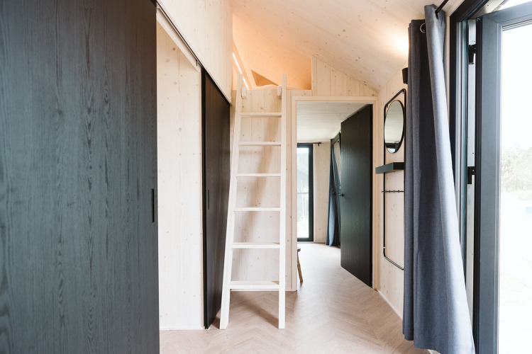Interior de una tiny house con escalera al altillo, paredes de madera clara y ventanas grandes en De Klepperstee, Países Bajos.