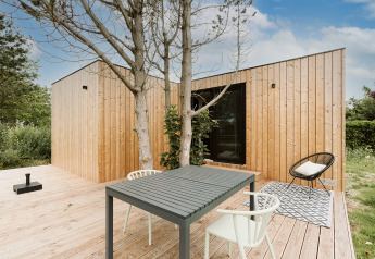 Modernes Tiny House mit Holzfassade, Terrasse, Esstisch und Stühlen, umgeben von Natur und Bäumen.
