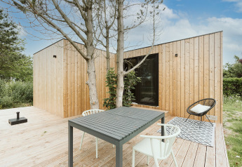 Casa pequeña moderna con fachada de madera, terraza exterior, mesa y sillas, rodeada de vegetación.