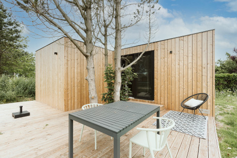 Tiny house moderne avec façade en bois, terrasse extérieure, table, chaises et vue sur la nature verdoyante.