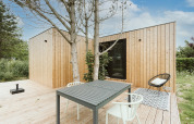 Casa pequeña moderna con fachada de madera, terraza exterior, mesa y sillas, rodeada de vegetación.