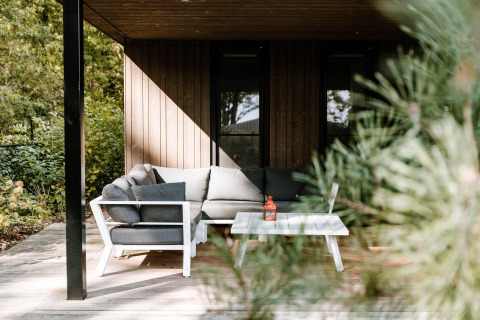 Acogedora terraza de lodge con sofá gris y mesa blanca, rodeada de vegetación y árboles iluminados por el sol.