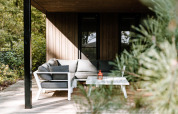 Hyggelig lodgeveranda med moderne grå sofa og hvidt bord, omgivet af frodige grønne træer i solen.