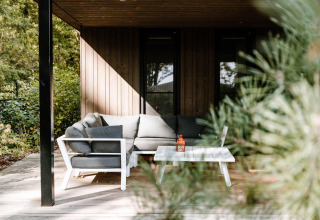 Knusse lodge veranda met grijze hoekzetel en witte tafel, omgeven door groen en bomen in het zonlicht.