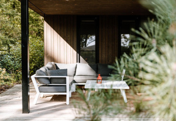Acogedora terraza de lodge con sofá gris y mesa blanca, rodeada de vegetación y árboles iluminados por el sol.