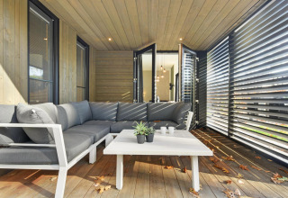 Moderne loungeområde på en lodge med grå sofa, hvidt bord og store vinduer, omgivet af træpaneler.