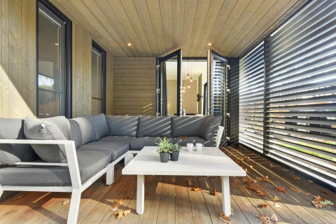 Moderne lodge veranda met grijze hoekbank, witte tafel, houten wanden en grote jaloezieën aan de zijkant.