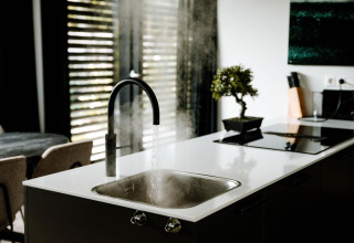 Évier moderne dans une cuisine de lodge, eau chaude qui coule, vapeur, plan de travail blanc et plante verte.