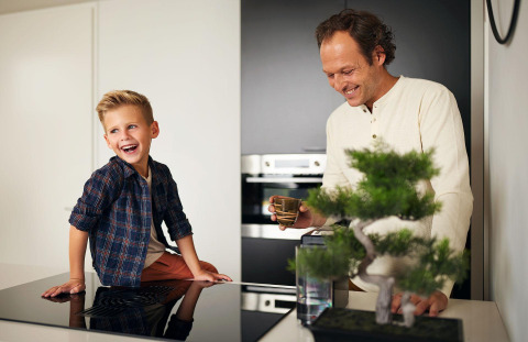 Father and son sharing laughter in the kitchen at Luxury Family Water Lodge, bonsai tree in foreground.