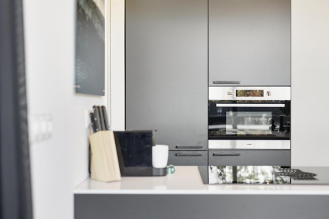 Cocina moderna en una cabaña con armarios grises, horno empotrado, bloque de cuchillos y encimera blanca.