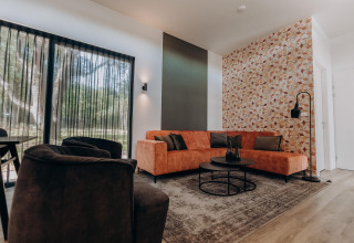 Moderne lodge woonkamer met grote ramen, oranje zetel, zwarte fauteuil en een opvallende wand.