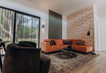 Moderne lodge woonkamer met grote ramen, oranje hoekbank, zwarte fauteuil en decoratieve wand.