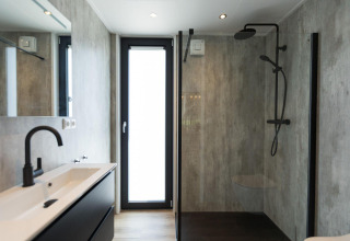 Salle de bain moderne dans une lodge avec douche en verre, robinetterie noire et grande porte lumineuse.