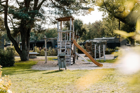 Parque infantil con torre de madera, tobogán y columpio junto a una cabaña rodeada de árboles y vegetación.
