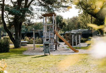 Parque infantil con torre de madera, tobogán y columpio junto a una cabaña rodeada de árboles y vegetación.