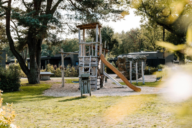 Playground with a wooden climbing tower, slide and swing at a lodge surrounded by trees and greenery.