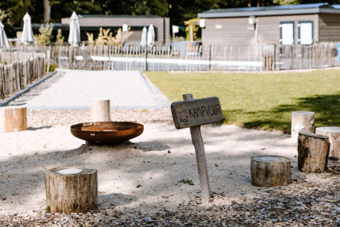 Bålplads ved lodge med træstubbe som siddepladser, skiltet 'KAMPVUUR' og baggrund med grøn græsplæne.