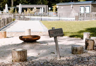 Kampvuurplek aan een lodge met boomstamstoelen, bord 'KAMPVUUR' en groen gras op de achtergrond.