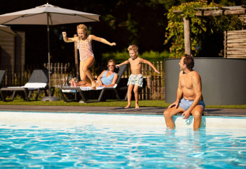 Dos niños saltan a una piscina mientras un hombre se sienta al borde y dos adultos descansan en tumbonas.