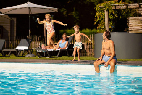 Two kids jump into a swimming pool as a man sits at the edge and two adults relax on loungers nearby.
