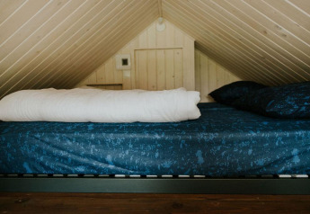 Letto nel soppalco con biancheria blu in Tiny House XL al Camping Si-Es-An, Paesi Bassi, sotto soffitto spiovente.