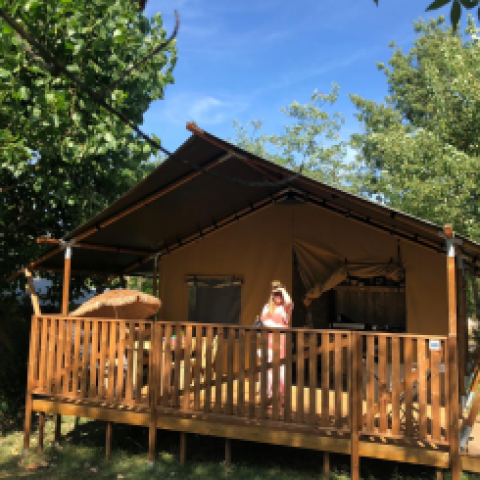 Tienda safari con instalaciones sanitarias en Camping Saint Amand, Francia, persona de pie en la terraza al sol.