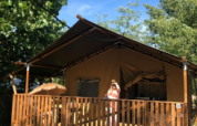Tienda safari con instalaciones sanitarias en Camping Saint Amand, Francia, persona de pie en la terraza al sol.