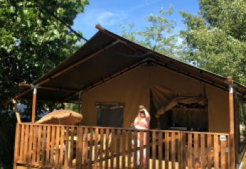 Tienda safari con instalaciones sanitarias en Camping Saint Amand, Francia, persona de pie en la terraza al sol.