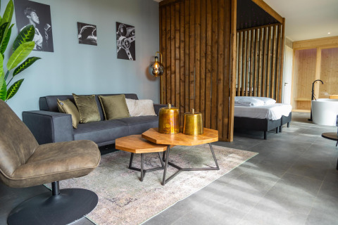 Sala moderna de lodge con sofá gris, mesa de madera y dormitorio visible en Modus Wellness Hoge Kempen.
