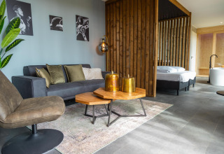 Zona giorno moderna in lodge con divano grigio, tavolino e camera da letto presso Modus Wellness Hoge Kempen.