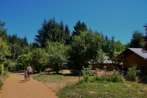 Día soleado en Châtel-Montagne, Francia, con personas caminando por un sendero rodeado de árboles y cabañas.