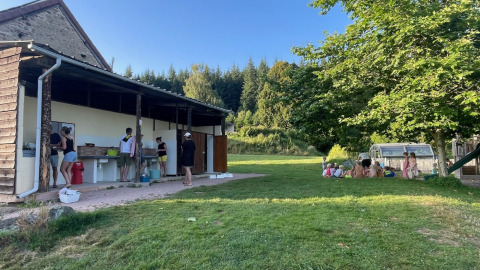 Personas lavando platos en una cocina exterior y niños jugando bajo árboles en un camping de Auvernia, Francia.