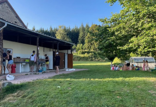 Folk vasker op i et udendørs køkken, mens børn leger under træer på en campingplads i Auvergne, Frankrig.