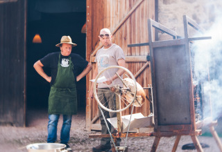 To mænd ved en udendørs grill, steger helstegt kød på Camping Domaine La Chabanne i Auvergne-Rhône-Alpes, Frankrig.