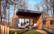 Modern wooden lodge with a covered deck and patio chairs, situated in a peaceful forest setting.