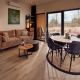 Moderne lodge-stue med beige sofa, rundt spisebord, store vinduer og udsigt til naturen udenfor.