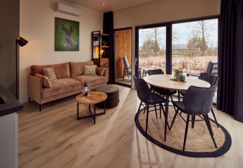 Séjour de lodge moderne avec canapé beige, table ronde, grandes fenêtres et vue sur la nature extérieure.
