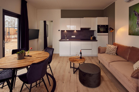 Acogedora sala y cocina de una cabaña moderna con mesa de madera, sofá, vino, plantas y cuadro mural.
