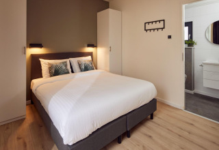 Modern lodge bedroom with a double bed, white bedding, wardrobe, wall lights, and en-suite bathroom view.