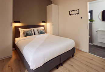 Moderne soveværelse i en lodge med dobbeltseng, hvidt sengetøj, garderobe og kig ind til badeværelse.