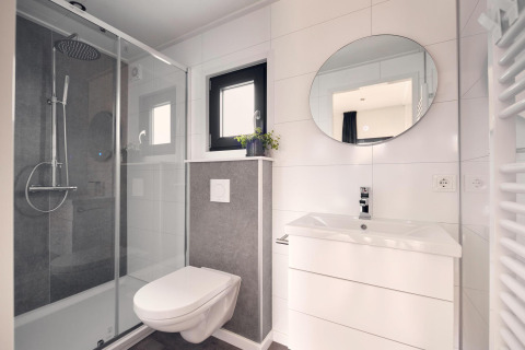 Modern lodge bathroom with walk-in glass shower, wall-mounted toilet, white vanity, and round mirror.