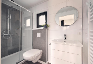 Baño moderno en un lodge con ducha acristalada, inodoro suspendido, lavabo blanco y espejo redondo.