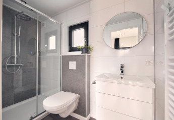 Modern lodge bathroom with walk-in glass shower, wall-mounted toilet, white vanity, and round mirror.