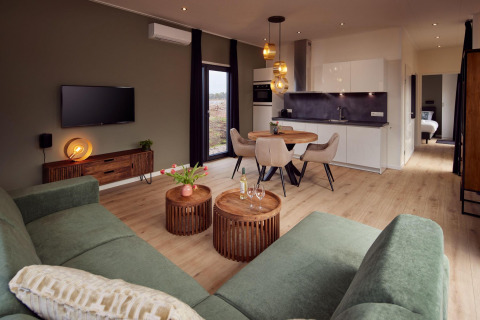 Modern lodge living room with green sofa, wooden tables, dining area, open kitchen, and bedroom in view.