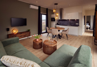 Moderne lodge stue med grøn sofa, træborde, spiseplads, åbent køkken og soveværelse i baggrunden.