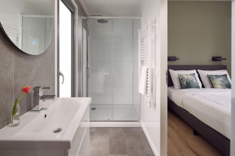 Interior moderno de lodge con baño luminoso, ducha amplia y vista al dormitorio con cama doble y cojines.