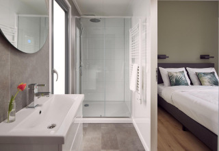 Interior moderno de lodge con baño luminoso, ducha amplia y vista al dormitorio con cama doble y cojines.
