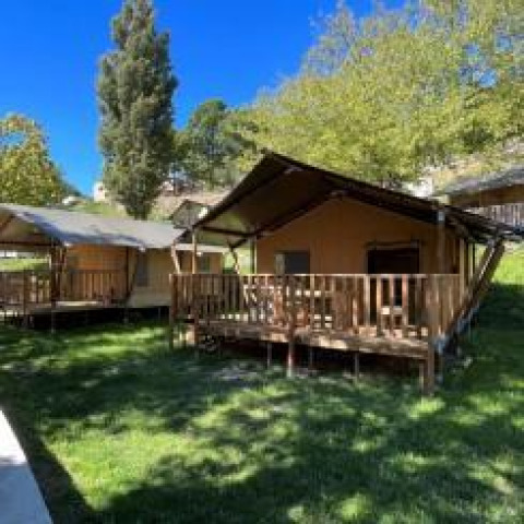 Safari tents with wooden decks set on green grass, surrounded by trees under a clear blue sky.