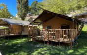 Safari tents with wooden decks set on green grass, surrounded by trees under a clear blue sky.